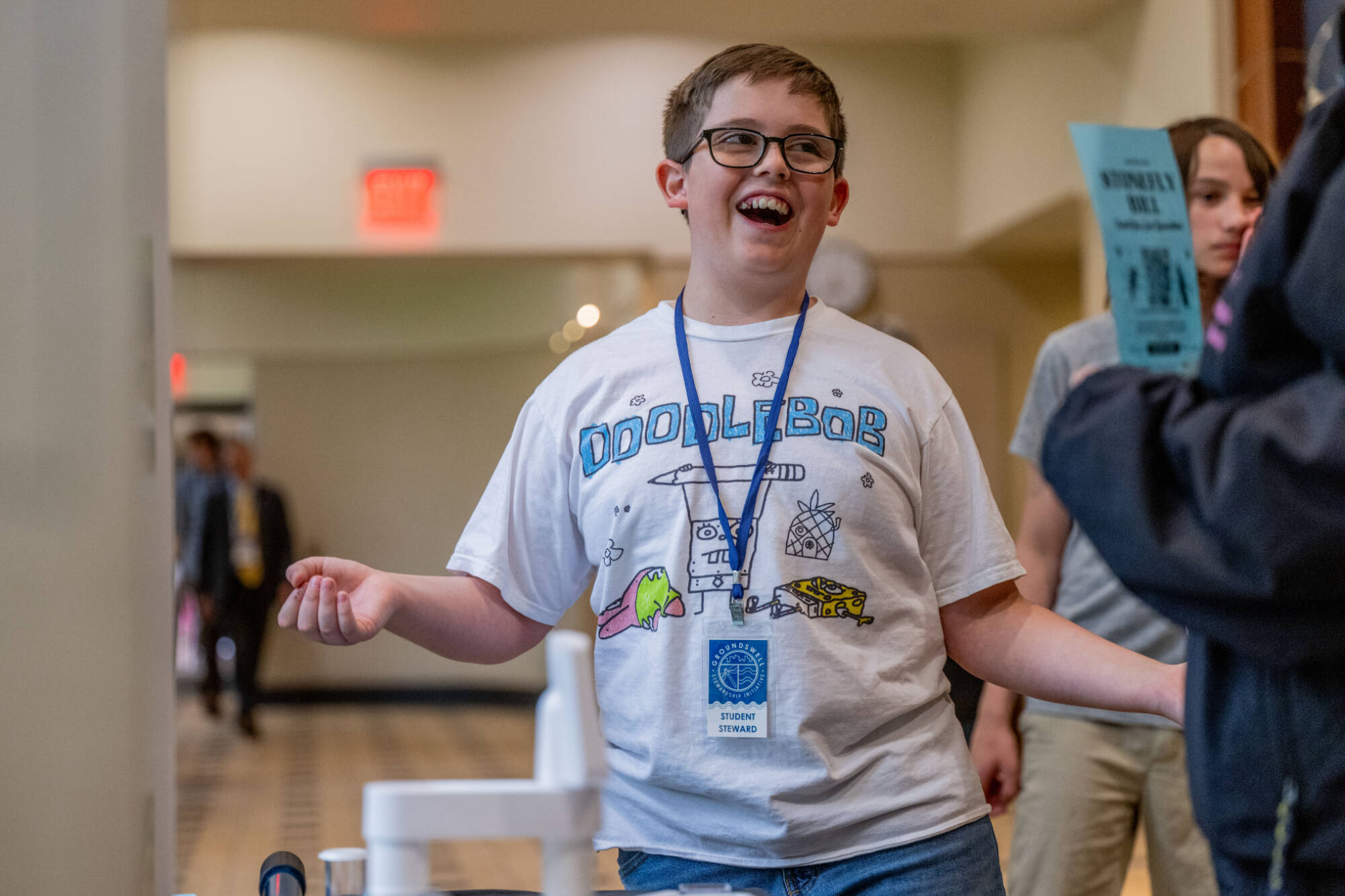 Laughing 5th-grade boy in a graphic T-shirt with a blue lanyard on, standing in a hallway of a large building
